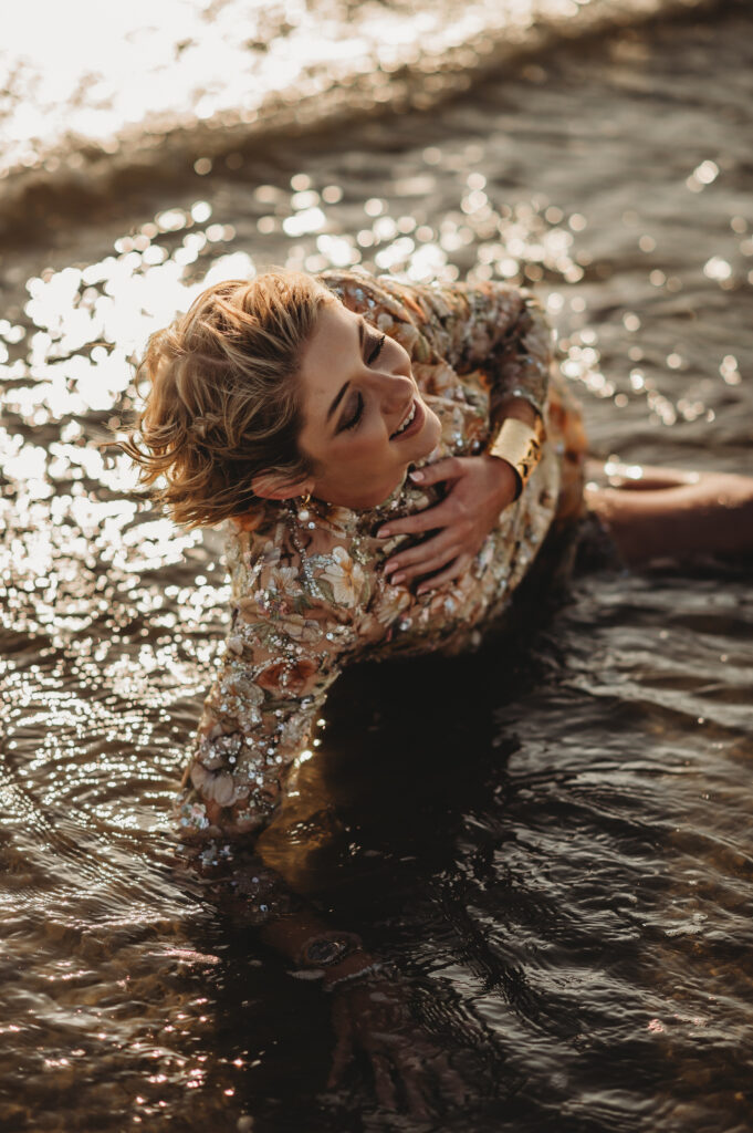 Woman in a shimmering sequin dress lying in the sea at sunset, surrounded by golden reflections, captured by Liberty Photography to symbolise rebirth, self-healing, and power after heartbreak.