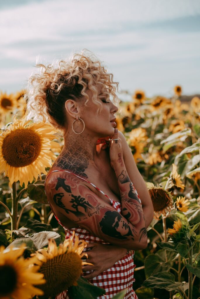 Blonde tattoed woman with curly hair standing in a field of sunflowers at sunset, captured by Liberty Photography during a summer empowerment shoot celebrating softness, confidence, and feminine energy.